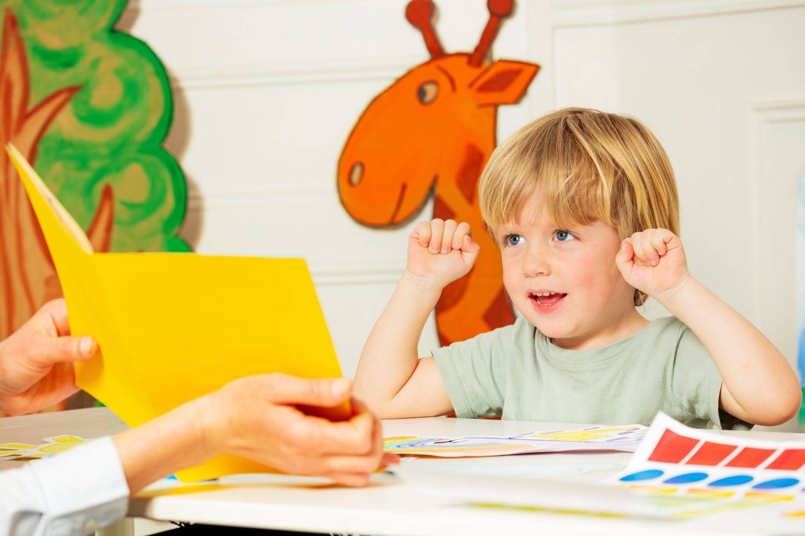 A young boy smiling at a table while an adult shows him a picture card